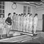 Row of naked boys stand on diving board at indoor pool